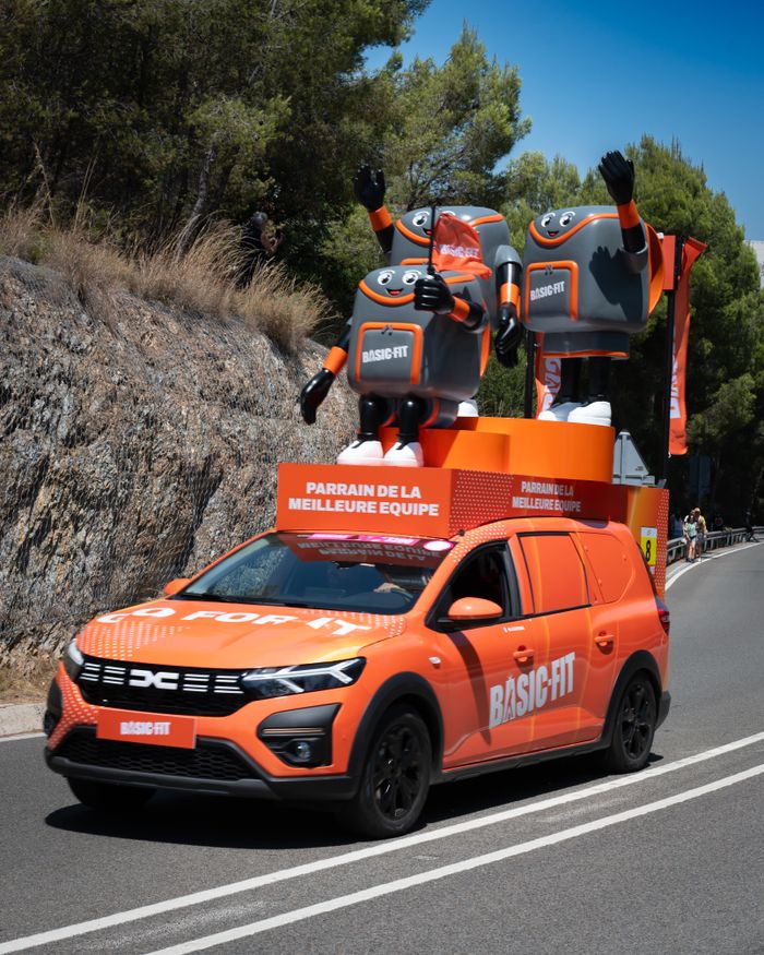 A bright orange car with mascots promotes a fitness brand, driving along a sunny road.